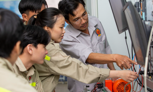 A male teacher at Ninh Thuan Vocational College is guiding a group of students in an electrical engineering practice session at the workshop. The whole group is focused on observing and operating equipment connected to a display screen.