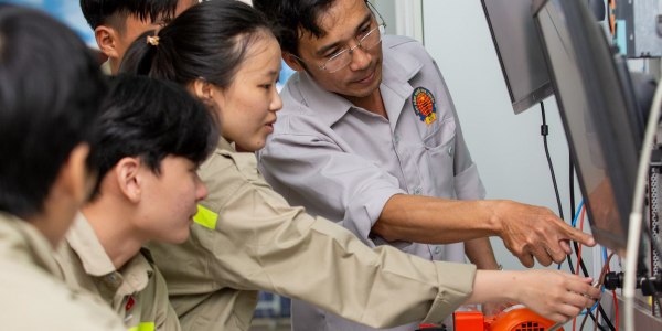A male teacher at Ninh Thuan Vocational College is guiding a group of students in an electrical engineering practice session at the workshop. The whole group is focused on observing and operating equipment connected to a display screen.