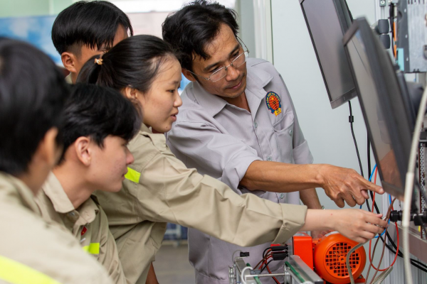 A male teacher at Ninh Thuan Vocational College is guiding a group of students in an electrical engineering practice session at the workshop. The whole group is focused on observing and operating equipment connected to a display screen.