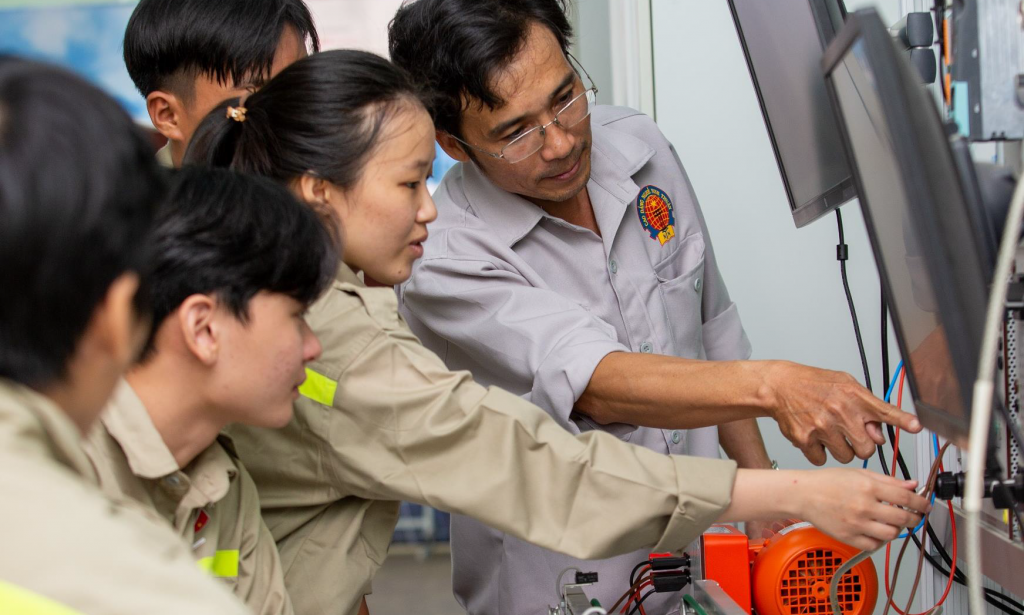 A male teacher at Ninh Thuan Vocational College is guiding a group of students in an electrical engineering practice session at the workshop. The whole group is focused on observing and operating equipment connected to a display screen.